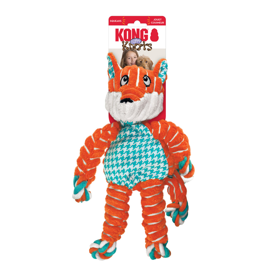 Toy in retail packaging. The packaging includes a red and white cardboard backing with the 'KONG Floppy Knots' logo. The plush toy resembles a fox with an orange and white color scheme, a soft fabric body, and twisted rope limbs. Its belly features a teal and white houndstooth pattern, and it has embroidered facial details. The toy's arms, legs, and tail have rope knots at the ends.