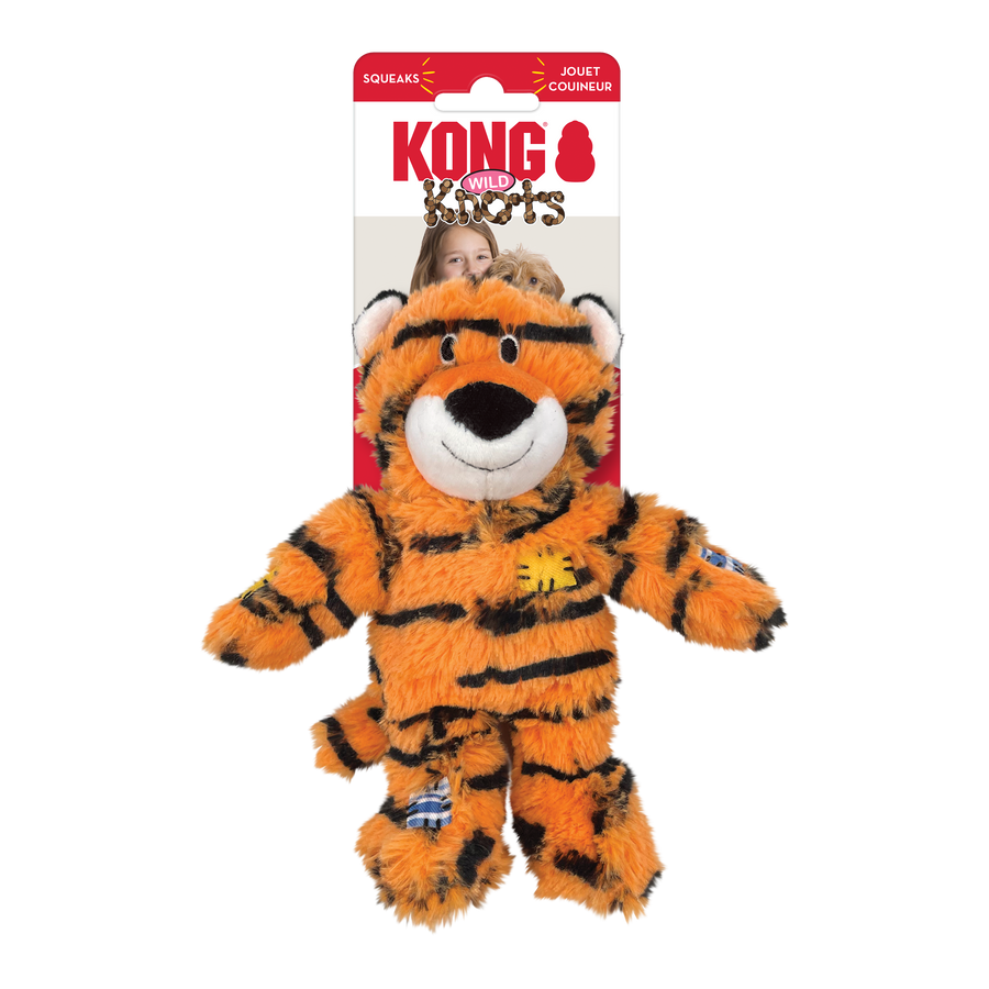 KONG Wild Knots Tiger Dog Toy in its original packaging, featuring a plush orange and black-striped tiger with a white muzzle and black nose. The toy has stitched black eyes, soft ears with white inner details, and a friendly smile. It includes decorative fabric patches in blue and yellow on its body and limbs. Designed with an internal knotted rope structure for durability, the plush toy also contains a squeaker. The toy's fluffy texture and lifelike tiger pattern make it visually appealing for pets.