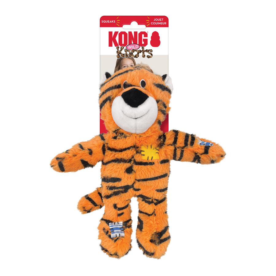 KONG Wild Knots Tiger Dog Toy in its packaging. The plush toy resembles a tiger with an orange body and black stripes. It has a soft, white muzzle, black nose, and a smiling expression with embroidered black eyes. The toy features fabric patches in yellow and blue stitched onto its body for a playful, rugged look. Its internal knotted rope structure enhances durability, while a squeaker inside adds interactive fun for pets. The toy is presented in KONG's signature red and white packaging.