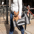 Bergan Booster Soft Pet Carrier, Side view of a woman is using the shoulder strap, with a small dog sitting inside the carrier. The dogs head is popped out of the carrier. 