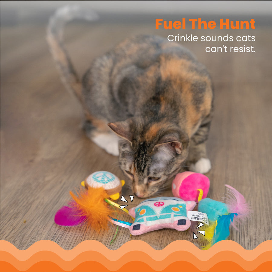 A calico cat sniffs a group of colorful toys on a wooden floor, including a plush van, mushroom, and tambourine toy with feathers. "Fuel the Hunt – Crinkle sounds cats can't resist."