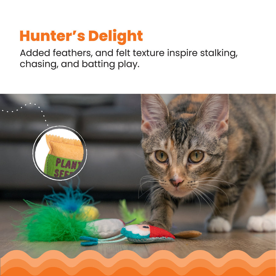 A tabby cat crouches near garden-themed cat toys on a wooden floor, including a gnome, bird, and flower with colorful feathers. An inset highlights a toy shaped like a seed packet labeled 'Plant Seeds.' Hunter’s Delight – Added feathers and felt texture inspire stalking, chasing, and batting play.