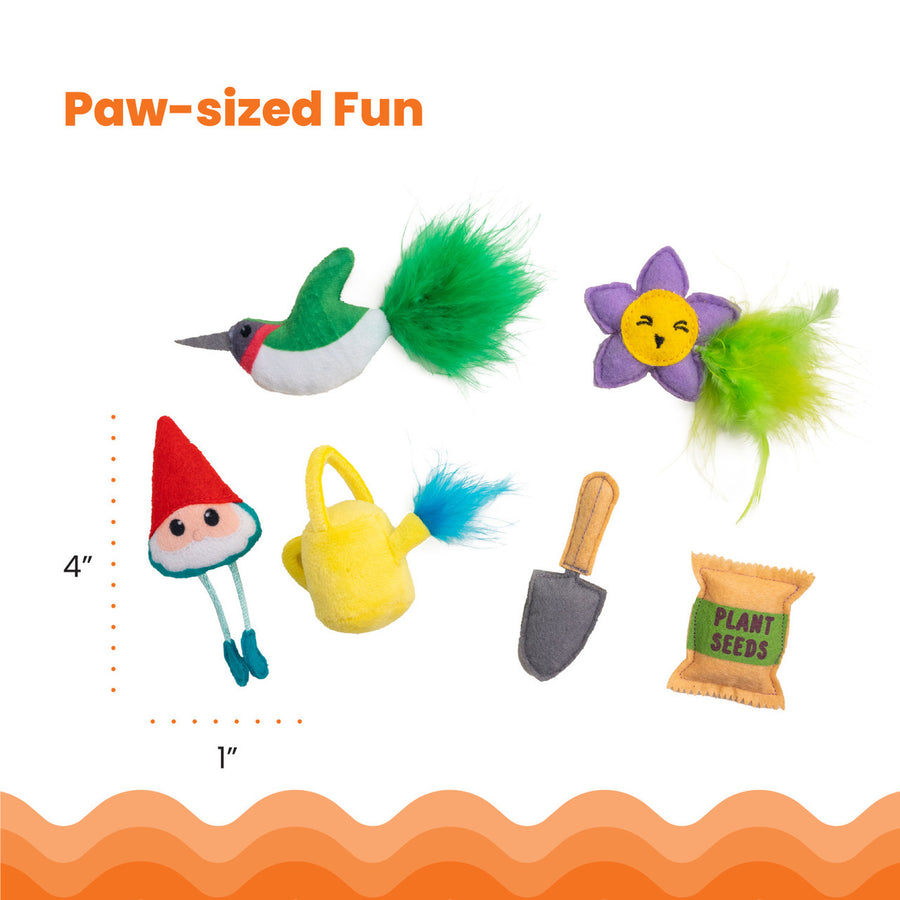 Six small garden-themed cat toys are arranged on a white background: a green hummingbird with a fluffy tail, a purple flower with green feathers, a red-hat garden gnome, a yellow watering can with blue feathers, a gray trowel, and a seed packet labeled 'Plant Seeds.' Dimensions shown are approximately 4 inches tall by 1 inch wide. Paw-sized Fun.