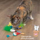 A calico cat sniffs a group of garden-themed cat toys on a wooden floor, including a hummingbird, flower, gnome, and trowel. In the corner, an illustrated cat peeks out of a flowerpot with a heart on it, next to the caption: 'Come back later… turning into a catalily.'