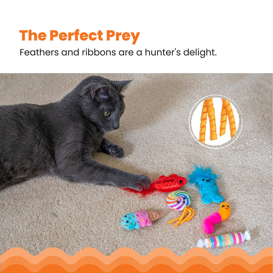 A gray cat lies on a carpeted floor, pawing at a red feather-tailed 'Sweet-ish' fish toy, surrounded by five other colorful candy-themed cat toys. An inset highlights curly orange ribbons. The Perfect Prey – Feathers and ribbons are a hunter's delight.