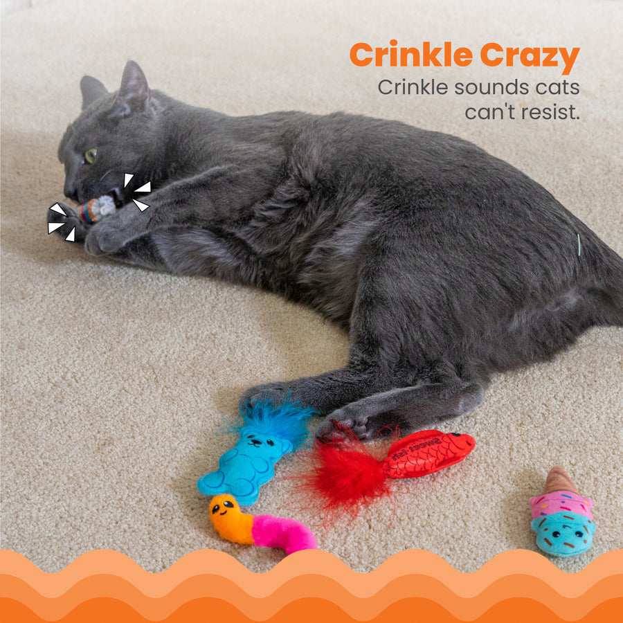A gray cat lies on a beige carpet, playfully biting a crinkly wrapped candy toy while surrounded by other colorful treats, including a blue gummy bear, a red feathered fish, an orange and pink worm, and an ice cream cone. Crinkle Crazy – Crinkle sounds cats can't resist.