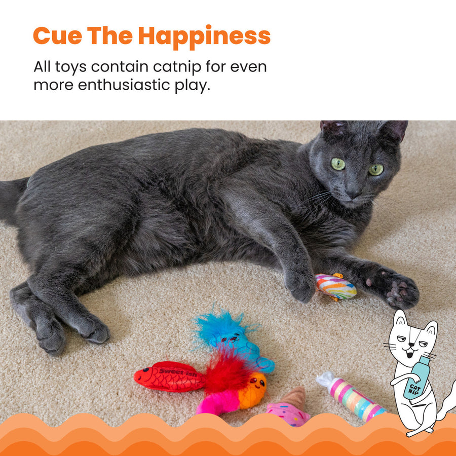 A gray cat lounges on a beige carpet, holding a rainbow lollipop cat toy while surrounded by other candy-themed toys, including a red fish, a gummy bear, a gummy worm, an ice cream cone, and a wrapped candy. Cue The Happiness – All toys contain catnip for even more enthusiastic play. An illustrated cat holding a catnip bottle appears in the corner.