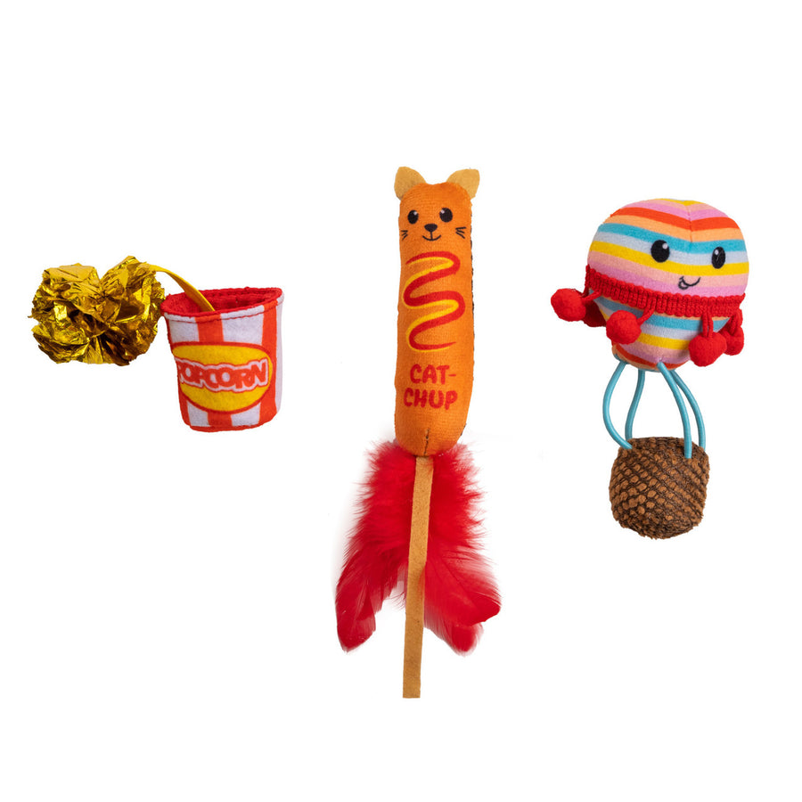 Three colorful cat toys with a playful party theme: a striped popcorn bucket with a shiny crinkle pom attached, an orange cat-shaped corn dog toy labeled 'CAT-CHUP' with red feathers at the bottom, and a round rainbow-striped toy with a smiling face, red pom-pom arms, blue legs, and a textured brown base.