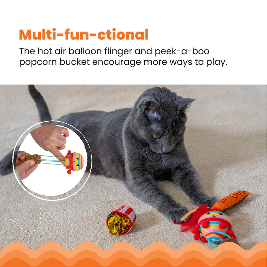 A gray cat plays with party-themed toys on a carpeted floor, including a corn dog toy with red feathers, a rainbow-striped balloon toy, and a popcorn bucket with shiny crinkle paper. An inset shows hands pulling and flinging the balloon toy. Multi-fun-ctional – The hot air balloon flinger and peek-a-boo popcorn bucket encourage more ways to play.