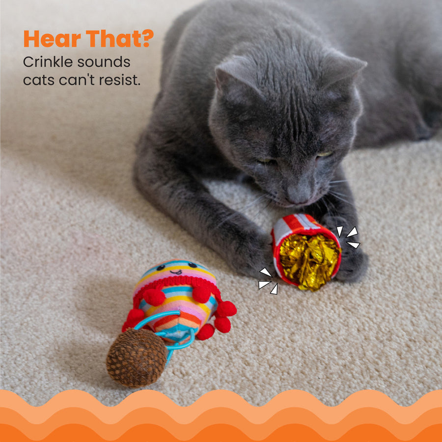 A gray cat paws at a red-and-white striped popcorn bucket cat toy filled with shiny gold crinkle paper, with a rainbow-striped hot air balloon toy nearby on the carpet. Hear That? – Crinkle sounds cats can't resist.