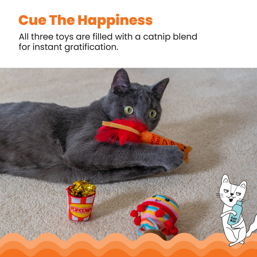 A gray cat lies on a beige carpet, clutching a corn dog-shaped cat toy with red feathers. Nearby are two other colorful toys: a popcorn bucket with shiny crinkle paper and a rainbow-striped hot air balloon. Cue The Happiness – All three toys are filled with a catnip blend for instant gratification. An illustrated cat holding a bottle labeled 'Catnip' appears in the corner.