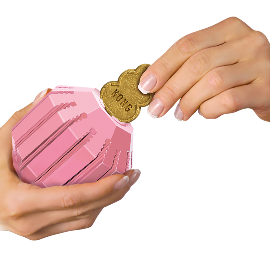 Close-up of two hands holding a pink KONG Puppy Activity Ball. One hand is inserting a brown KONG-shaped treat into the toy's grooves, demonstrating its treat-dispensing feature.