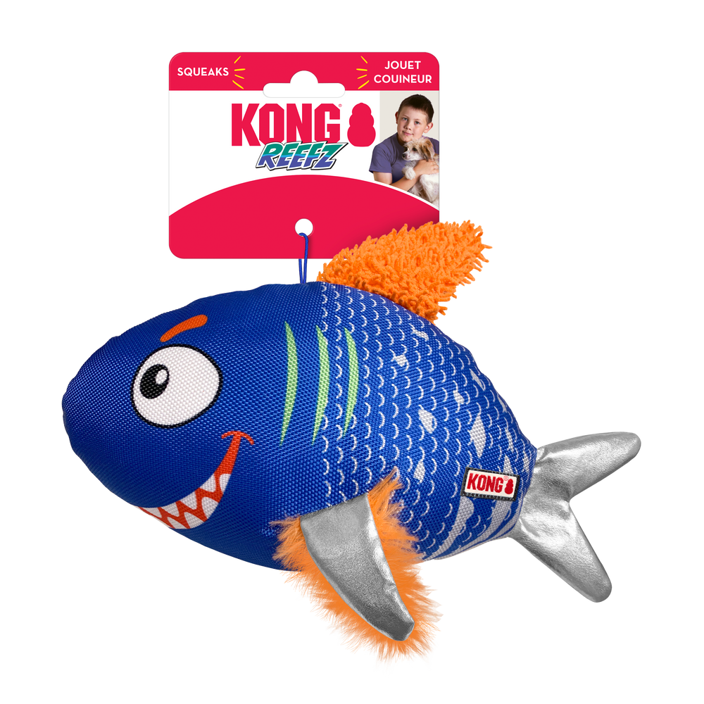 A KONG Reefz dog toy in its original packaging. The toy is shaped like a cartoonish fish with a blue textured fabric body, white scale patterns, and an expressive face with large eyes and a toothy grin. It has silver fabric fins, an orange plush tail, and orange fuzzy side fins. The packaging is red and white, displaying the KONG logo, the word 'Reefz' in a blue and green stylized font. The packaging also highlights that the toy squeaks.