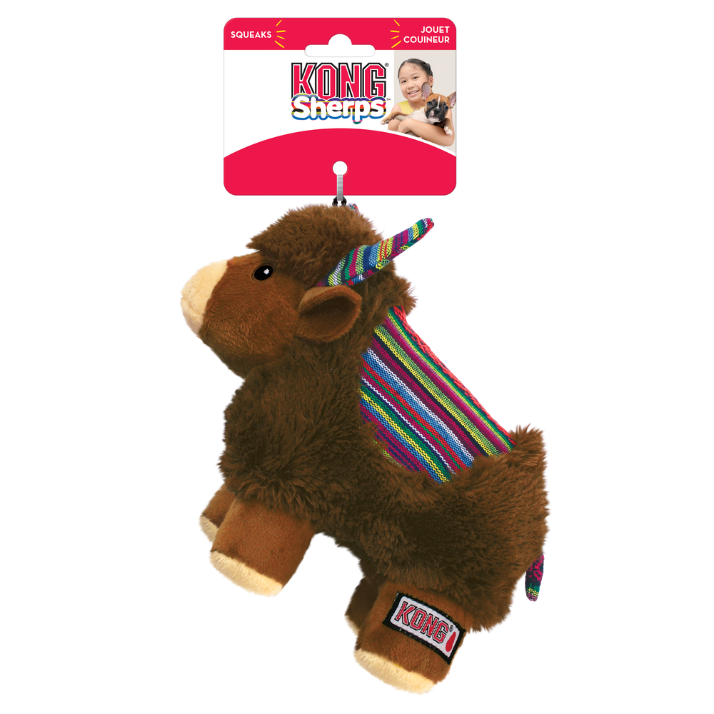  KONG Sherps yak plush dog toy in its original packaging. The toy has a soft, brown furry body with a beige muzzle and embroidered eyes. Its ears and curved horns feature a colorful woven fabric with bright striped patterns, which match the saddle-like fabric on its back. The yak stands on beige plush legs with white hooves and has a short, fluffy tail. The packaging is red and white, displaying the KONG logo, the word 'Sherps' in a playful blue font, also highlights that the toy squeaks.