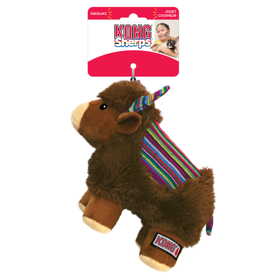  KONG Sherps yak plush dog toy in its original packaging. The toy has a soft, brown furry body with a beige muzzle and embroidered eyes. Its ears and curved horns feature a colorful woven fabric with bright striped patterns, which match the saddle-like fabric on its back. The yak stands on beige plush legs with white hooves and has a short, fluffy tail. The packaging is red and white, displaying the KONG logo, the word 'Sherps' in a playful blue font, also highlights that the toy squeaks.