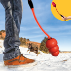 A person using the KONG Signature KONG Throw dog toy in a sandy outdoor setting. The image focuses on the thrower, which is red and black, launching a classic red KONG-shaped rubber toy. The person's legs and brown sneakers are visible in the foreground. In the background, a brown and black dog excitedly runs toward the toy. The landscape features sandy ground, dry grass, and rocky formations under a blue sky. In the top right corner, a yellow circular inset shows a close-up of a hand gripping thrower.