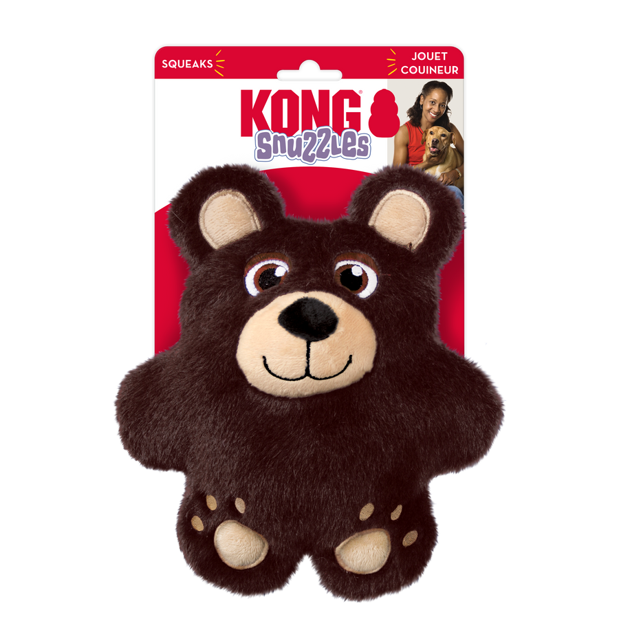 A KONG Snuzzles Bear plush dog toy in its original packaging. The toy is a soft, round, brown bear with a fuzzy texture. It features embroidered eyes, a beige muzzle with a black plush nose, and a stitched smiling mouth. The bear has rounded ears with beige inner linings and small embroidered paw prints on its feet. The packaging is red and white, displaying the KONG logo, the word 'Snuzzles'. The packaging also highlights that the to that the toy squeaks.