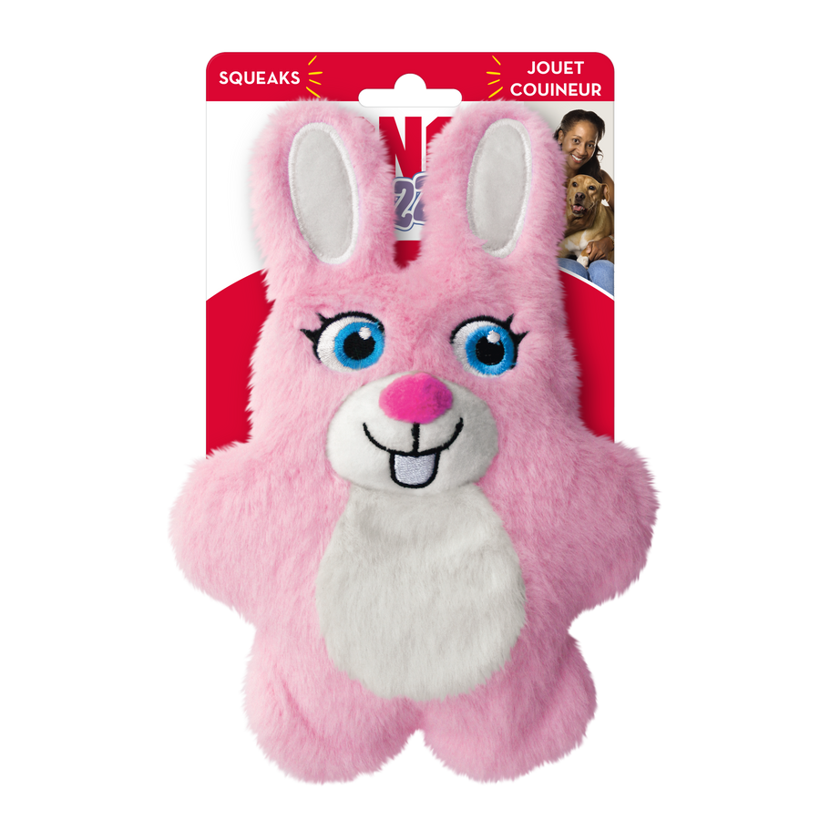 A KONG Snuzzles Kiddos Bunny plush dog toy in its original packaging. The toy is a soft, round, pink bunny with a fuzzy texture. It features embroidered blue eyes with long lashes, a white muzzle with a pink plush nose, a stitched black smile, and two front teeth. The bunny has long, upright ears with white inner linings and a white fluffy patch on its chest. The packaging is red and white, displaying the KONG logo, the word 'Snuzzles'. The packaging also highlights that the toy squeaks.