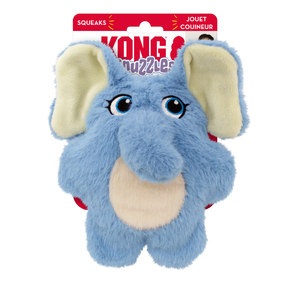 A KONG Snuzzles Kiddos Elephant plush dog toy in its original packaging. The toy is a soft, round, light blue elephant with a fuzzy texture. It features embroidered blue eyes with long lashes, a plush trunk, and a beige oval patch on its belly. The elephant has large, floppy ears with cream-colored inner linings. The packaging is red and white, displaying the KONG logo, the word 'Snuzzles' in a playful purple font. The packaging also highlights that the toy squeaks.