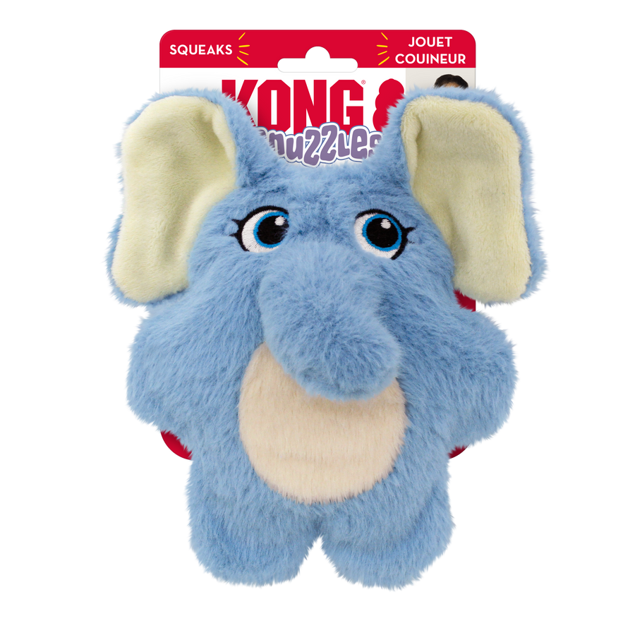 A KONG Snuzzles Kiddos Elephant plush dog toy in its original packaging. The toy is a soft, round, light blue elephant with a fuzzy texture. It features embroidered blue eyes with long lashes, a plush trunk, and a beige oval patch on its belly. The elephant has large, floppy ears with cream-colored inner linings. The packaging is red and white, displaying the KONG logo, the word 'Snuzzles' in a playful purple font. The packaging also highlights that the toy squeaks.