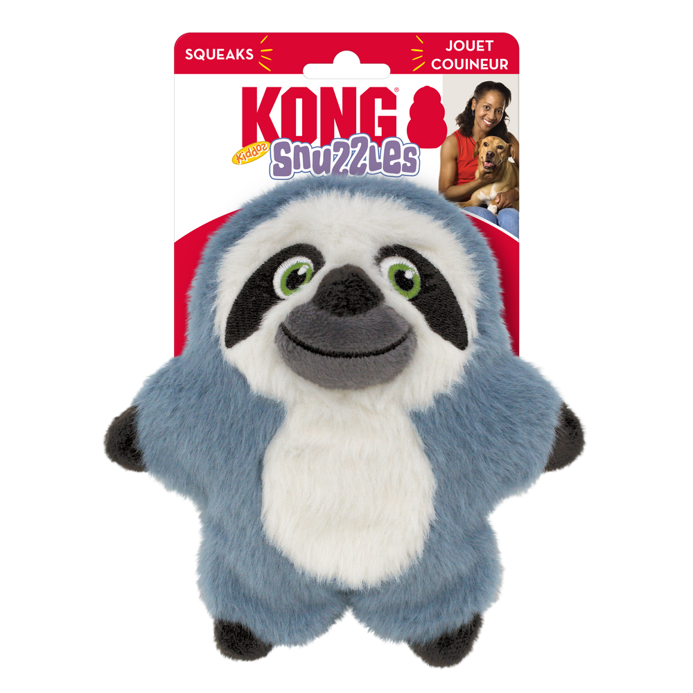 A KONG Snuzzles Kiddos Sloth plush dog toy in its original packaging. The toy is a soft, round, blue-gray sloth with a fuzzy texture. It has a smiling face with embroidered green eyes, a black nose, and a white face framed by dark brown patches. The plush toy also features a white oval patch on its belly and small black paws. The packaging is red and white, displaying the KONG logo, the word 'Snuzzles' in a playful purple font. The packaging also highlights that the toy squeaks.