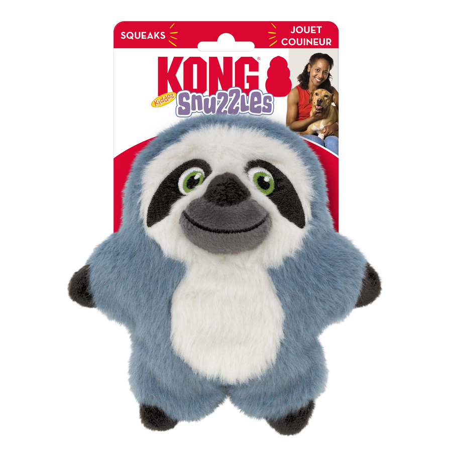 A KONG Snuzzles Kiddos Sloth plush dog toy in its original packaging. The toy is a soft, round, blue-gray sloth with a fuzzy texture. It has a smiling face with embroidered green eyes, a black nose, and a white face framed by dark brown patches. The plush toy also features a white oval patch on its belly and small black paws. The packaging is red and white, displaying the KONG logo, the word 'Snuzzles' in a playful purple font. The packaging also highlights that the toy squeaks.