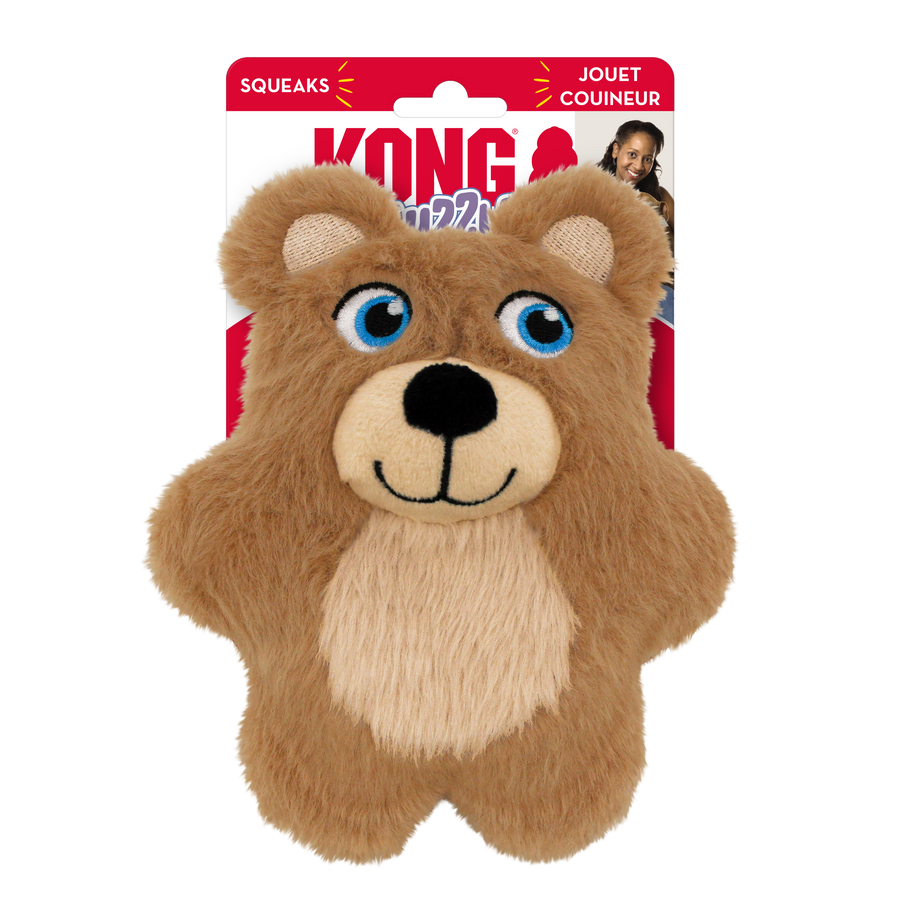 A KONG Snuzzles Kiddos Teddy plush dog toy in its packaging. The toy is a soft, round, light brown teddy bear with a fuzzy texture. It has embroidered blue eyes, a black nose, and a stitched smiling mouth. A beige patch is present on its belly, contrasting with the brown fur. The toy is designed for dogs, offering a cuddly and interactive play experience with a built-in squeaker. The background features the KONG branding with red and white colors.