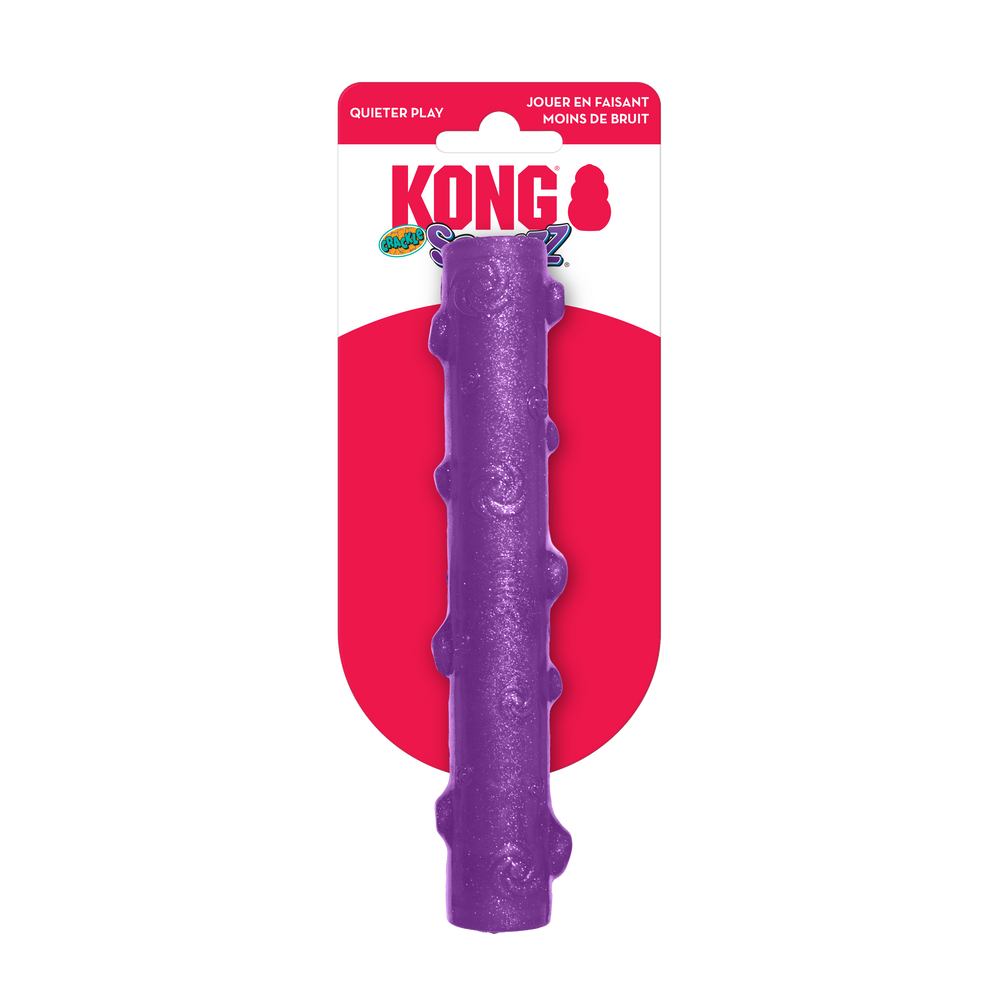 A KONG Squeezz Crackle Stick dog toy in its original packaging. The toy is a long, cylindrical stick-shaped chew toy in vibrant purple with a slightly textured surface featuring raised bumps. It is designed for interactive play and features a crackling sound for added engagement. The packaging has a red and white KONG-branded card with the words 'Quieter Play' in the upper left corner, indicating its noise-reducing design.