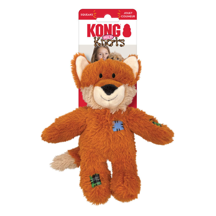 The KONG Wild Knots Fox Dog Toy, a plush orange fox with a friendly face, soft fur, and a white muzzle. It has floppy arms and legs with knotted rope inside for added durability. The toy features decorative stitched patches on its body and limbs. The packaging in the background is red and white, displaying the KONG Wild Knots logo.