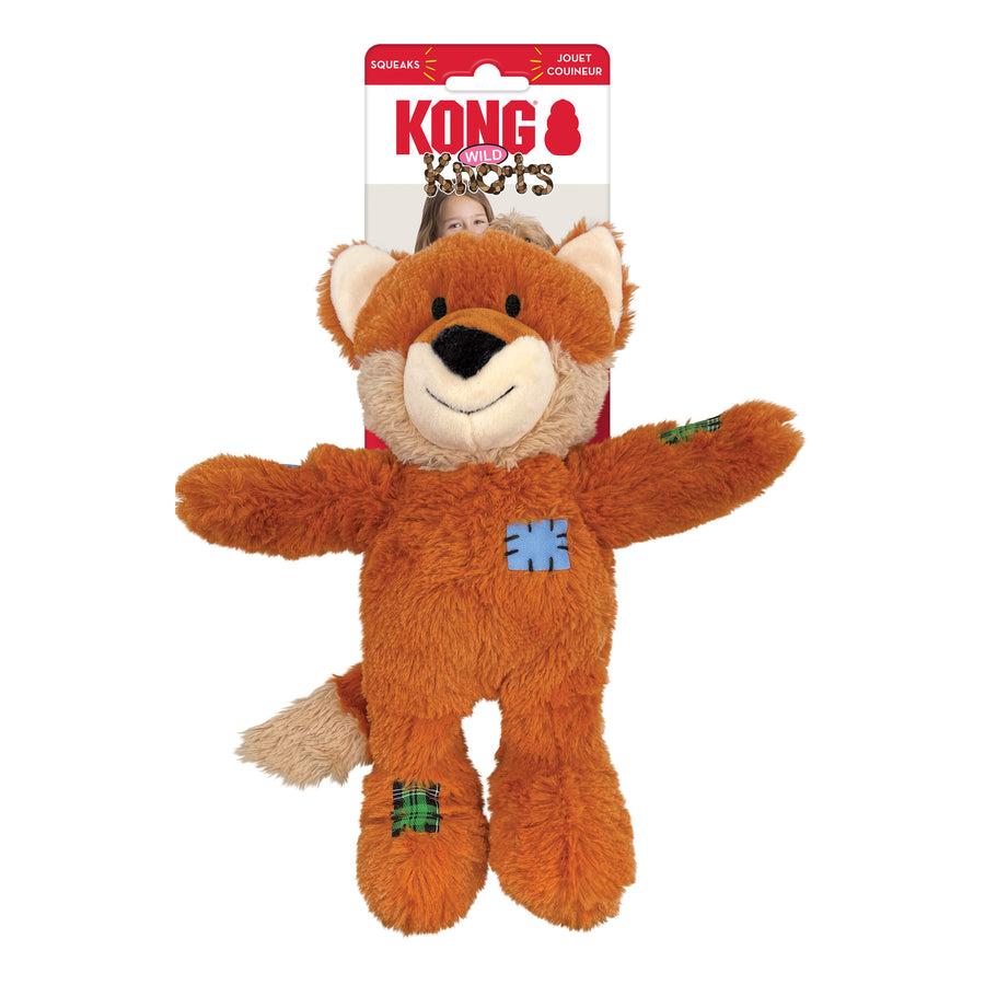 KONG Wild Knots Fox Dog Toy in its original packaging. The plush fox toy has soft orange fur with a white muzzle, inner ears, and tail tip. It features a smiling expression and stitched patch details in blue and green plaid on its body and limbs. Designed for durability, the toy includes an internal knotted rope structure while maintaining a soft exterior. The packaging has a red and white KONG branding with a young girl and her dog in the background.