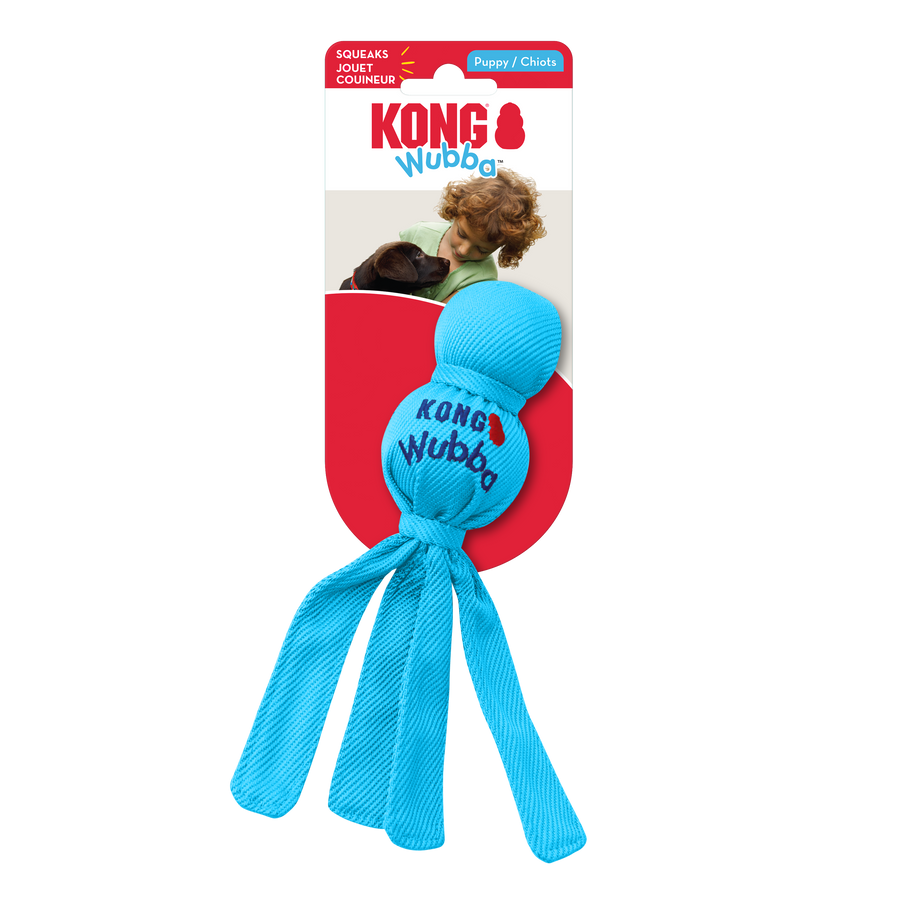 A KONG Wubba Puppy dog toy in its original packaging. The toy is bright blue with a fabric-covered ball-shaped top and long fabric tails hanging below. The packaging has a red and white background, featuring the KONG logo, the word 'Wubba' in blue. The packaging also highlights that the toy squeaks and is designed for puppies
