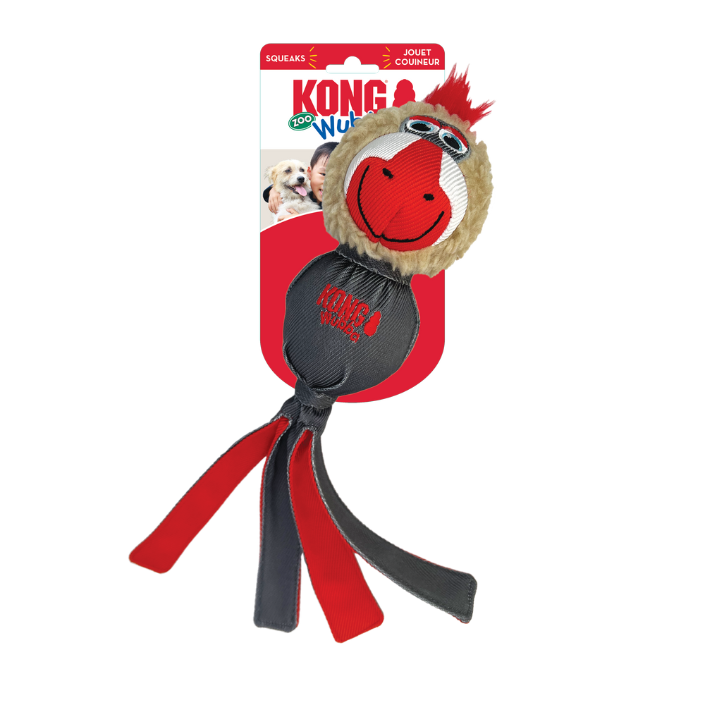 A KONG Wubba Zoo Mandrill Dog Toy in its original packaging. The toy has a round, plush head resembling a mandrill, with a tan furry face, large embroidered eyes, a red and white snout, and a tuft of red fur on top. The body is made of durable dark gray fabric, with the 'KONG Wubba' logo printed in red. Multiple long fabric tails in red and gray extend from the bottom, designed for interactive play such as tugging and shaking.