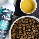 Nulo Omega 3-6-9 Oil Blend Supplement for Dogs, a bottle of the oil supplement next to a bowl of dry dog food and a small bowl of the oil for mixing