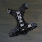 Wilderdog black dog harness laid flat on dark wood surface, showing chevron reflective strip, mesh padding, quick-release buckles, and D-ring.