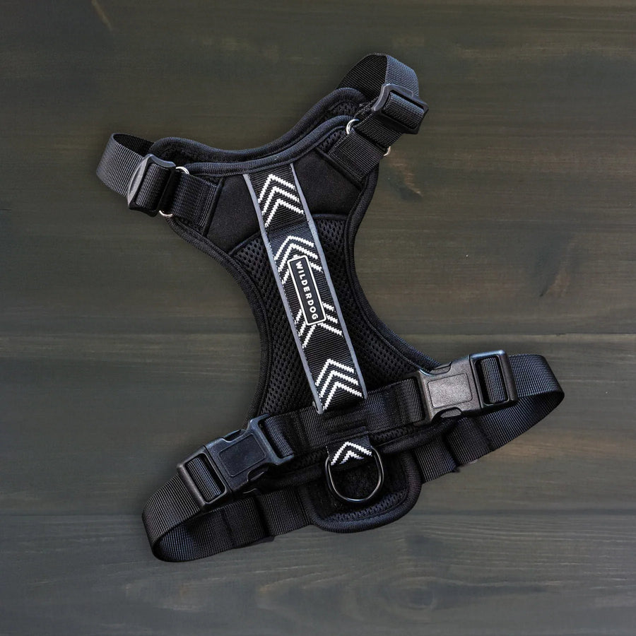 Wilderdog black dog harness laid flat on dark wood surface, showing chevron reflective strip, mesh padding, quick-release buckles, and D-ring.