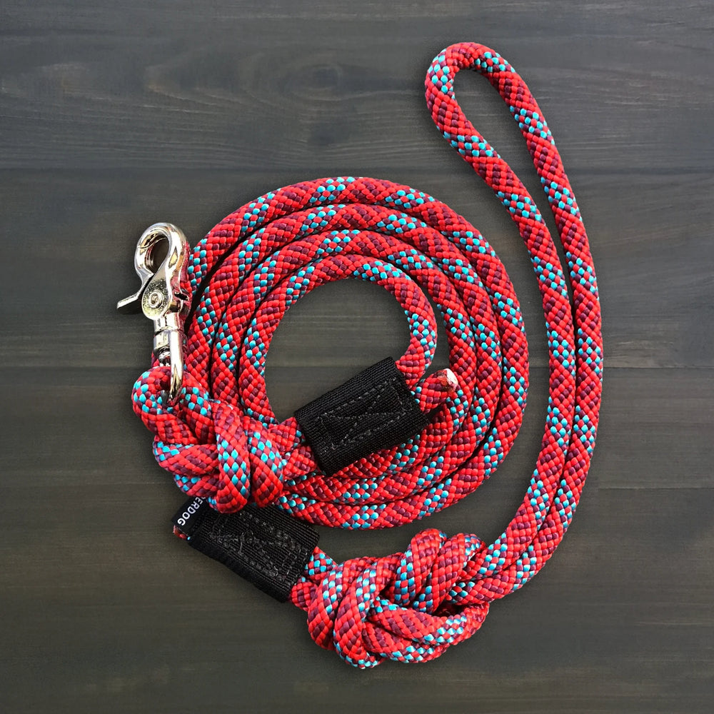 Wilderdog Quick Clip Dog Rope Leash coiled on dark wood surface, red and teal braided rope with silver snap clip and black Wilderdog branded grip.