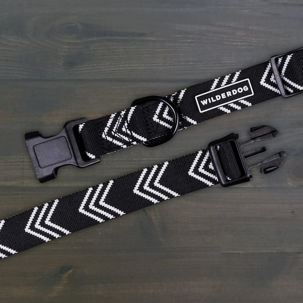 Wilderdog Orca ultralight dog collar laid flat on dark wood surface, showing black webbing with white chevron arrow pattern, Wilderdog logo patch, black D-ring, and quick-release buckle.