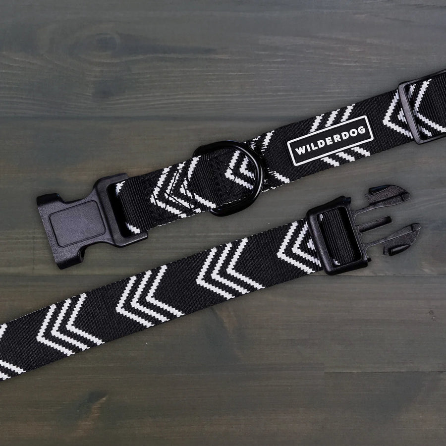 Wilderdog Orca ultralight dog collar laid flat on dark wood surface, showing black webbing with white chevron arrow pattern, Wilderdog logo patch, black D-ring, and quick-release buckle.