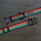 Wilderdog Trout ultralight dog collar laid flat on dark wood surface, showing black webbing with gradient rainbow trout scale pattern in red, orange, teal, and green with dark spot detailing, Wilderdog logo patch, black D-ring, and quick-release buckle.