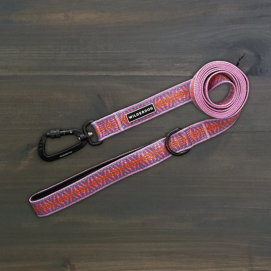 Wilderdog Ultralight Dog Leash in Lilac laid flat on dark wood surface, pink and orange speckled woven webbing with black Wilderdog carabiner clip, D-ring, and branded label.