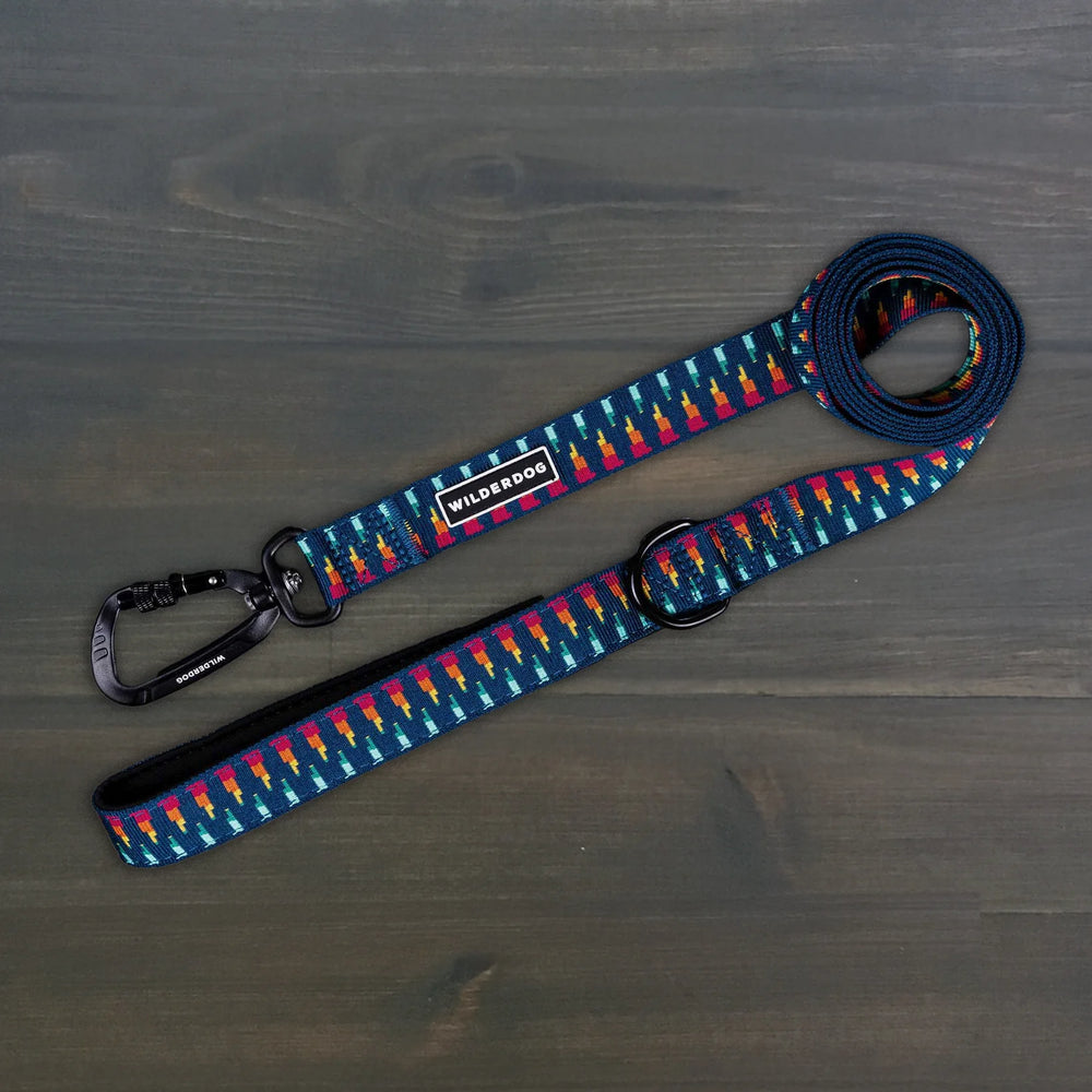 Wilderdog Ultralight Dog Leash in Marina colorway laid flat on dark wood surface, navy blue webbing with multicolor geometric pattern, black Wilderdog carabiner clip, D-ring, and branded label.