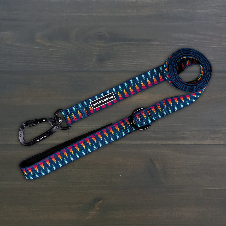 Wilderdog Ultralight Dog Leash in Marina colorway laid flat on dark wood surface, navy blue webbing with multicolor geometric pattern, black Wilderdog carabiner clip, D-ring, and branded label.