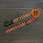 Wilderdog Ultralight Dog Leash in Sienna laid flat on dark wood surface, orange, red, and teal striped woven pattern with black Wilderdog carabiner clip, D-ring, and branded label.
