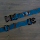 Wilderdog Pacific Blue waterproof dog collar laid flat on dark wood surface, showing teal blue coated webbing with subtle topo map line pattern, Wilderdog logo patch, black D-ring, and black quick-release buckle.