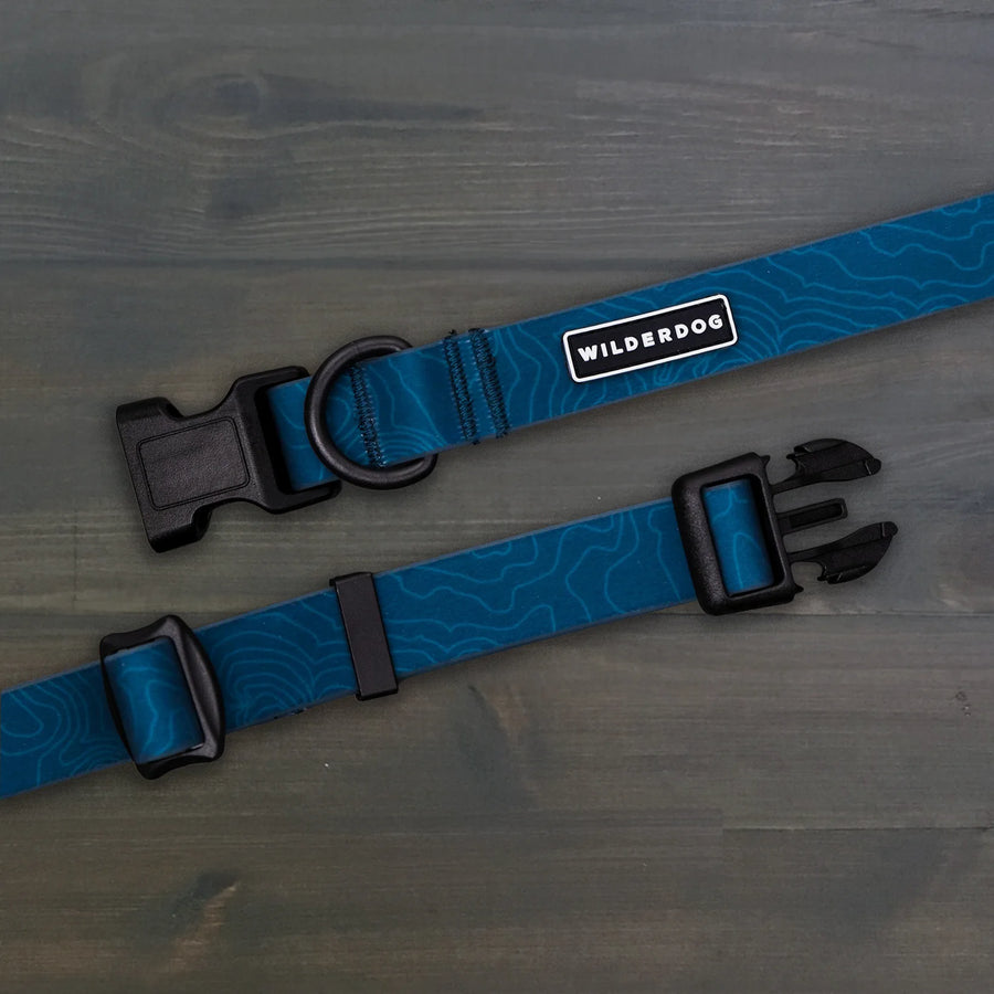 Wilderdog Pacific Blue waterproof dog collar laid flat on dark wood surface, showing teal blue coated webbing with subtle topo map line pattern, Wilderdog logo patch, black D-ring, and black quick-release buckle.