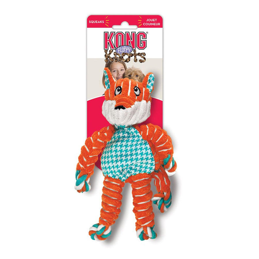 Toy in retail packaging. The packaging includes a red and white cardboard backing with the 'KONG Floppy Knots' logo. The plush toy resembles a fox with an orange and white color scheme, a soft fabric body, and twisted rope limbs. Its belly features a teal and white houndstooth pattern, and it has embroidered facial details. The toy's arms, legs, and tail have rope knots at the ends. 
