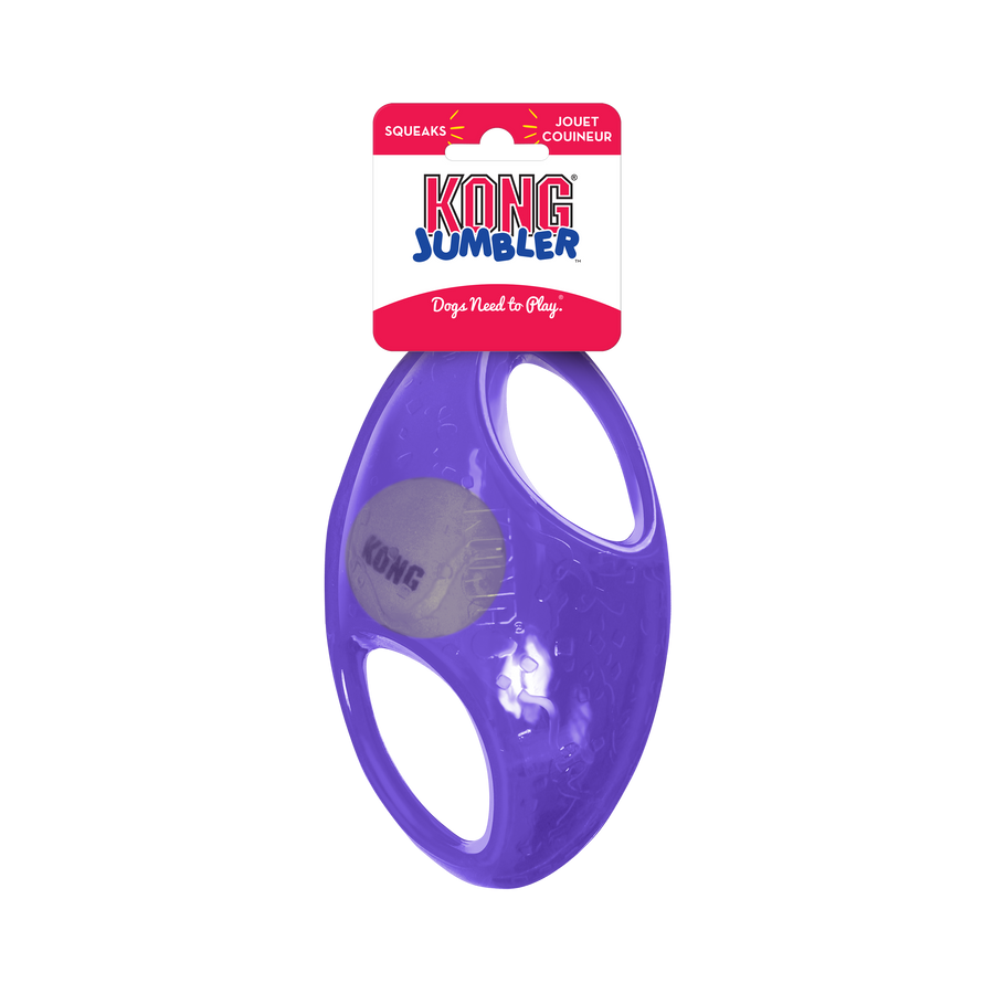 A purple KONG Jumbler Football Dog Toy in its packaging. The toy features a durable, transparent rubber outer shell with two built-in handles for easy gripping. Inside the toy, a smaller KONG-branded tennis ball is visible. The packaging at the top is red and white, displaying the KONG Jumbler logo along with the words 'Dogs Need to Play' and indicating that the toy squeaks.